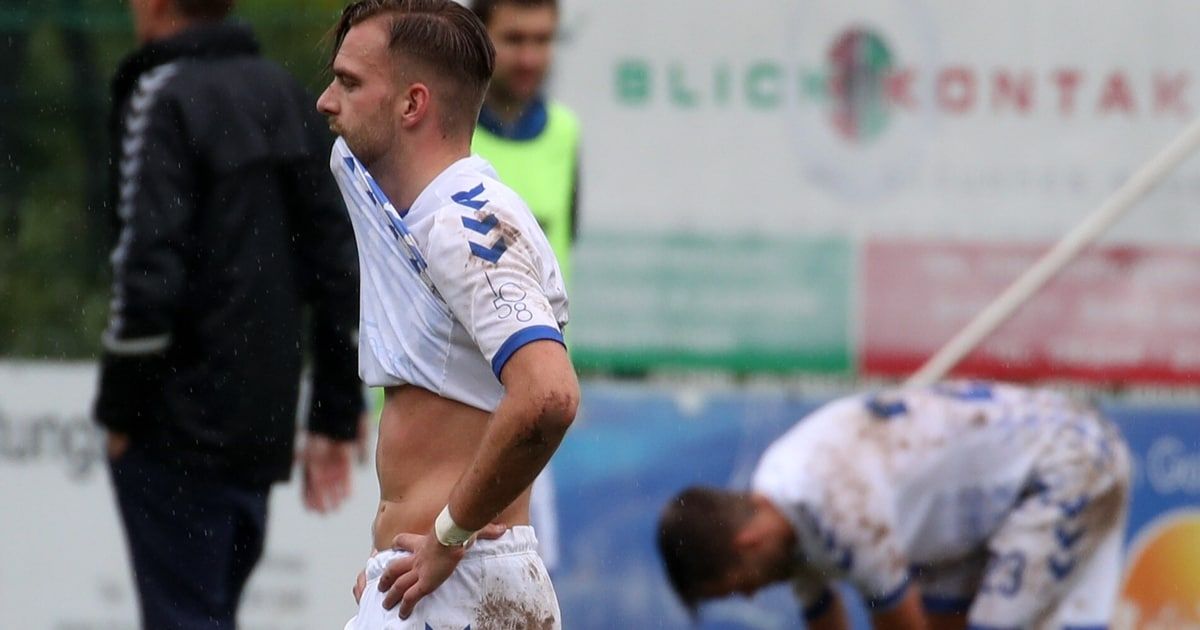 A soccer player in a white jersey with blue trim, number 30, stands on a field, his shirt pulled up to his neck, looking to the side. In the background, another player bends over.