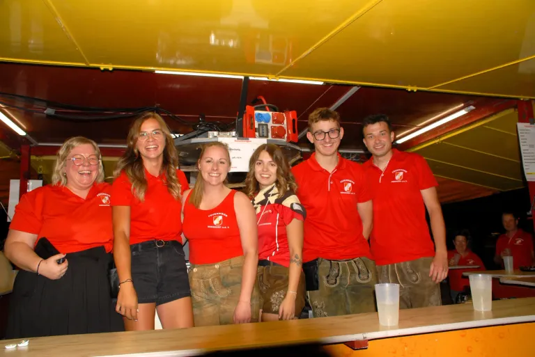 A group of five people in red shirts and shorts are standing together in a room, smiling for a photo. They are wearing glasses, bracelets, and one has a tattoo. A desk with a cup is in front of them.