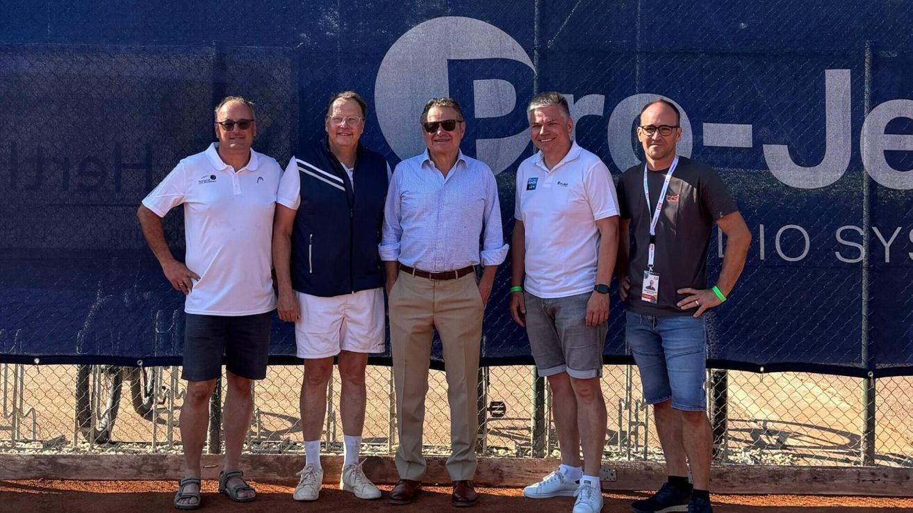 Five men stand together in front of a blue backdrop with a logo. They are all smiling and appear to be posing for a photo.