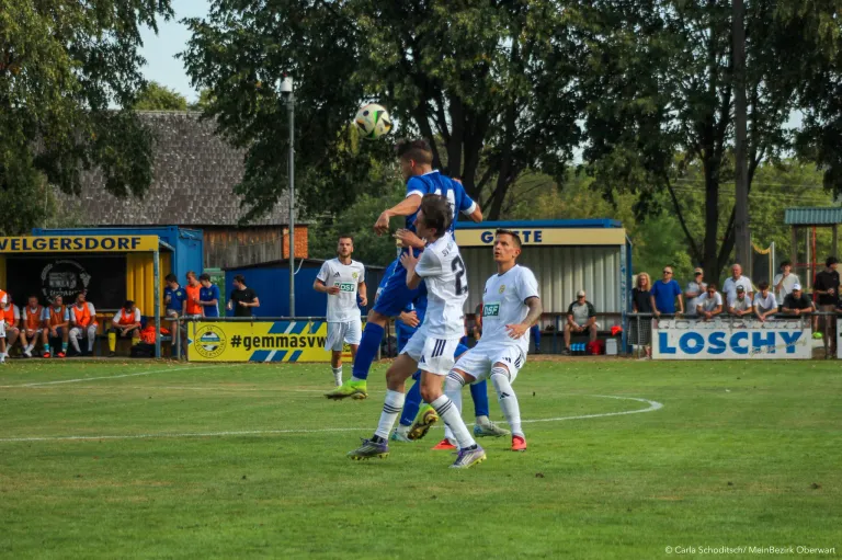 Soccer players on a field, one player in blue jumps to head the ball, while two others in white try to block him.