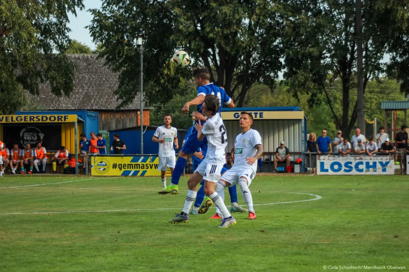 Soccer players on a field, one player in blue jumps to head the ball, while two others in white try to block him.