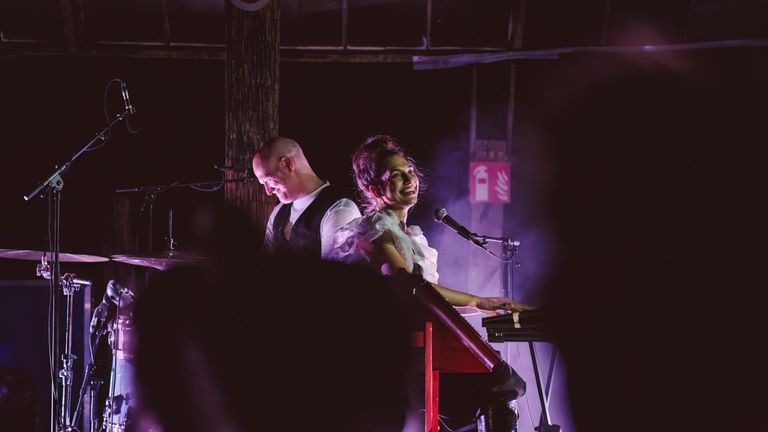A woman plays a keyboard while a man plays drums on stage under purple lighting.