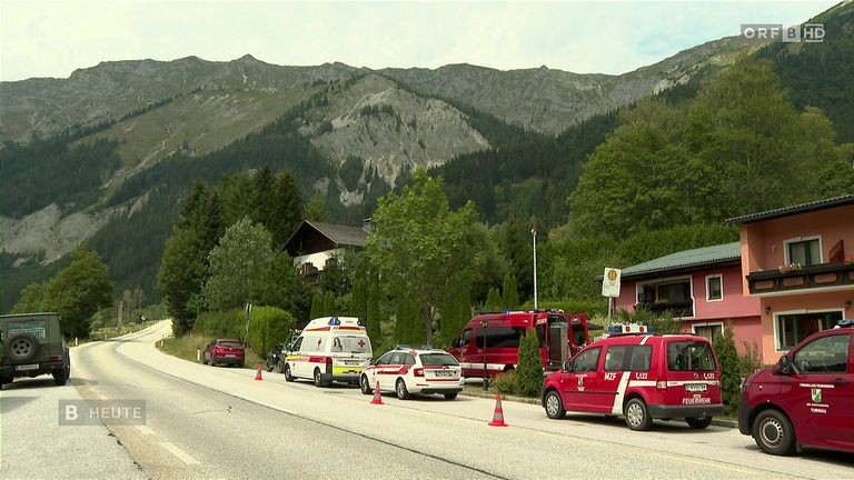 Emergency vehicles are parked on the side of a road with mountains in the background. Trees and bushes line the roadside. A building with a sign is visible on the right.