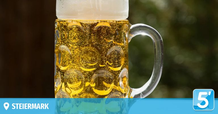 A close-up view of a glass mug filled with golden beer, showing numerous bubbles rising to the top. The background is slightly blurred, suggesting a sunny outdoor setting.