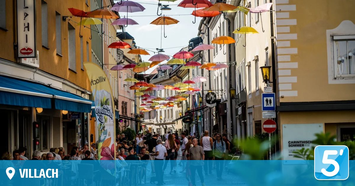 Eine geschäftige Stadtstraße mit bunten Regenschirmen darüber, Menschen, die gehen und in Outdoor-Cafés sitzen, und Gebäude auf beiden Seiten.