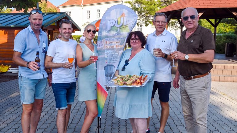 Four adults stand in a line, smiling and holding drinks, with one woman holding a tray of food. They are in front of a banner and a building.