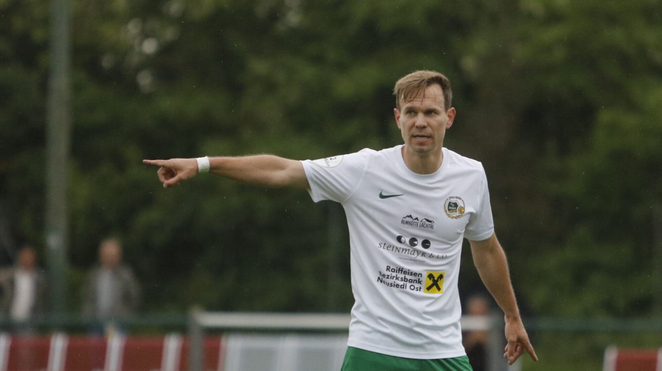 Ein Mann in einem weißen T-Shirt mit mehreren Logos, einschließlich Nike, zeigt nach links. Er trägt grüne Shorts und ein weißes Armband. Der Hintergrund ist unscharf mit Bäumen.