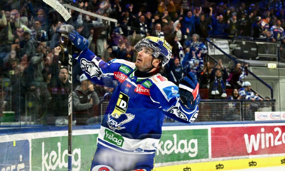 Ein Eishockeyspieler feiert ein Tor vor einer jubelnden Menge. Er trägt ein blaues Trikot mit verschiedenen Sponsorenlogos und hält einen Eishockeyschläger. Die Eisbahn ist von einer Glasbarriere mit Werbung umgeben.