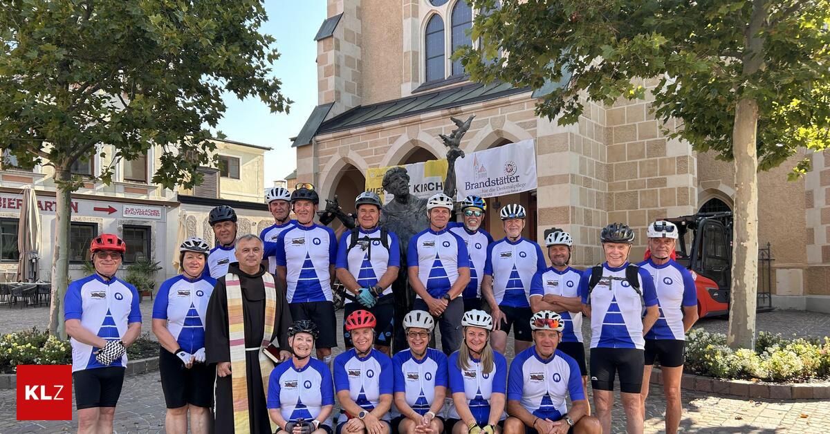 Eine Gruppe von Radfahrern in passenden blauen und weißen Uniformen posiert für ein Foto vor einer Kirche. Ein Priester steht unter ihnen, und eine Statue befindet sich hinter ihnen.