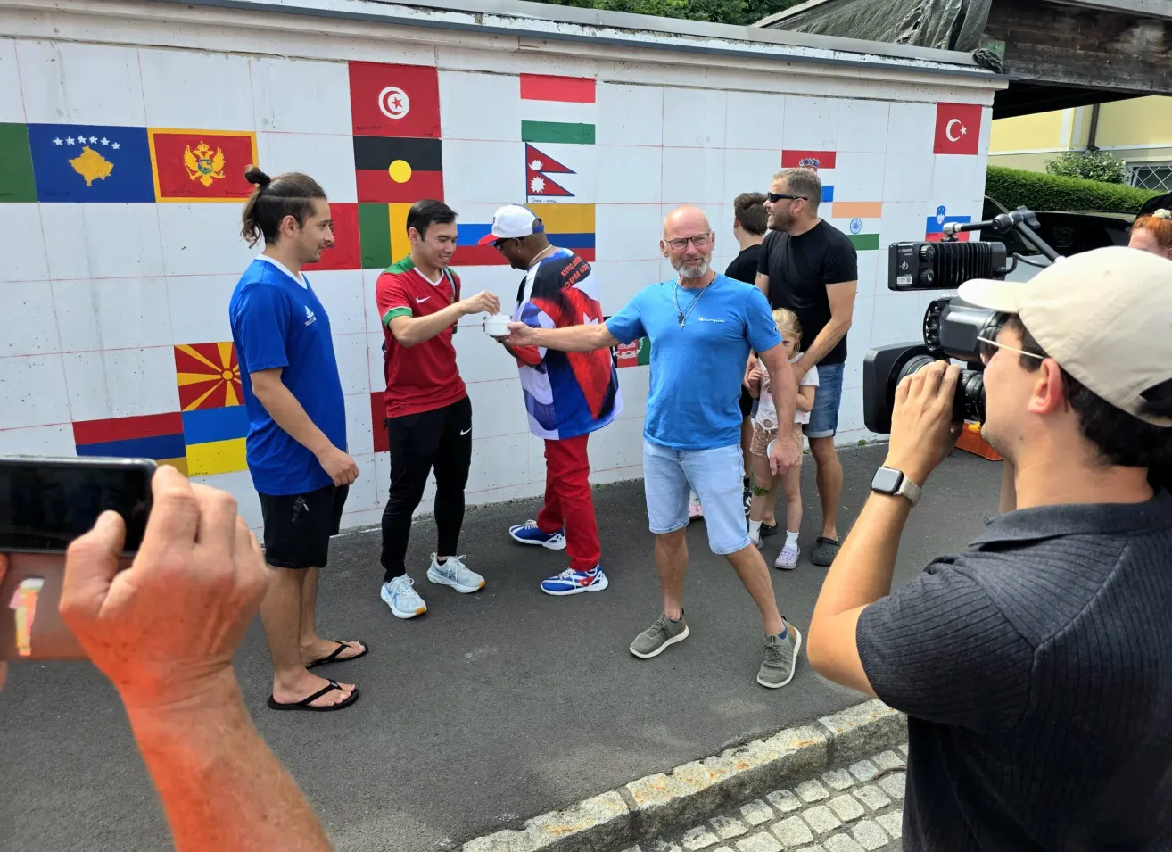Eine Gruppe von Männern in verschiedenfarbigen Trikots und Turnschuhen steht vor einer Wand mit Flaggen. Ein Mann reicht einem anderen eine Tasse. Ein Fotograf hält den Moment fest.