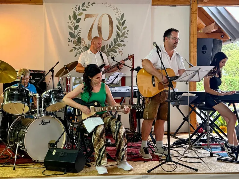 Eine Band tritt auf der Bühne auf, hinter ihnen ein Banner mit der Aufschrift 70. Die Frau spielt Gitarre, während die beiden Männer Gitarre und Schlagzeug spielen.