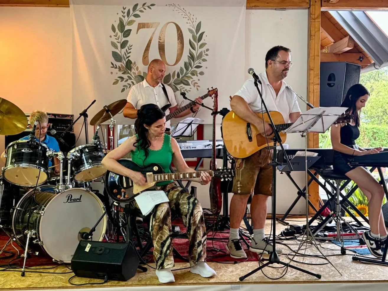 A band is performing on stage with a banner behind them that says 70. The woman is playing the guitar, while the two men are playing the guitar and drums.
