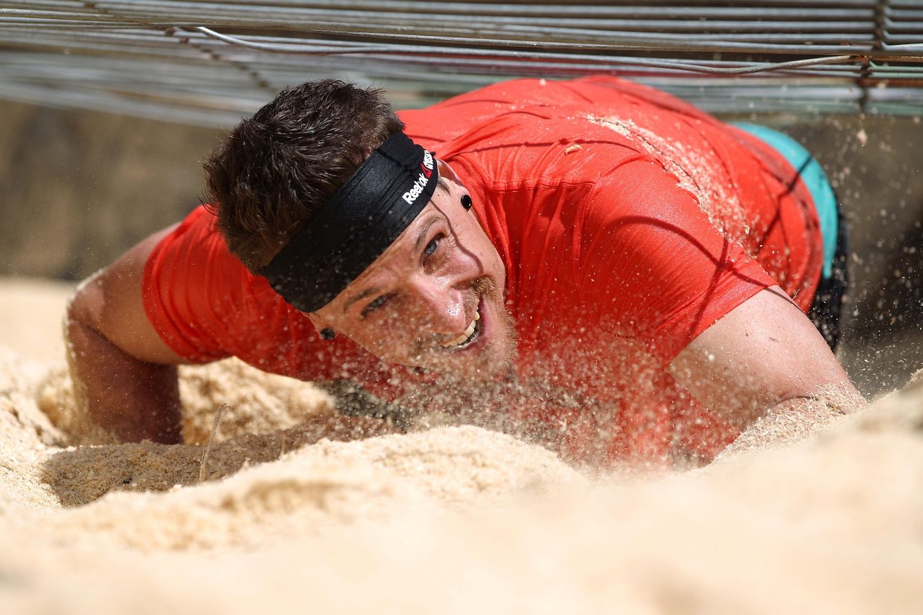 Ein Mann in einem roten Shirt und Kopfband kriecht durch einen sandigen Hindernisparcours, lächelt und ist mit Sand bedeckt.