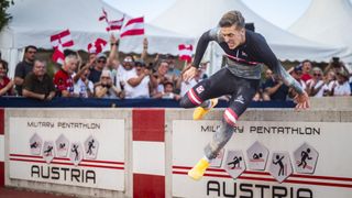 Ein Mann in einem schwarzen und roten Anzug springt über eine Barriere bei einem Triathlon-Event, während eine Menge hinter ihm jubelt.