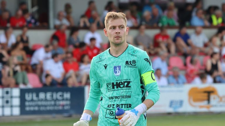 Ein Fußballtorwart in einem grünen Trikot steht auf dem Spielfeld und hält eine Wasserflasche in der rechten Hand. Dahinter ist eine verschwommene Menge von Zuschauern, die das Spiel verfolgen.