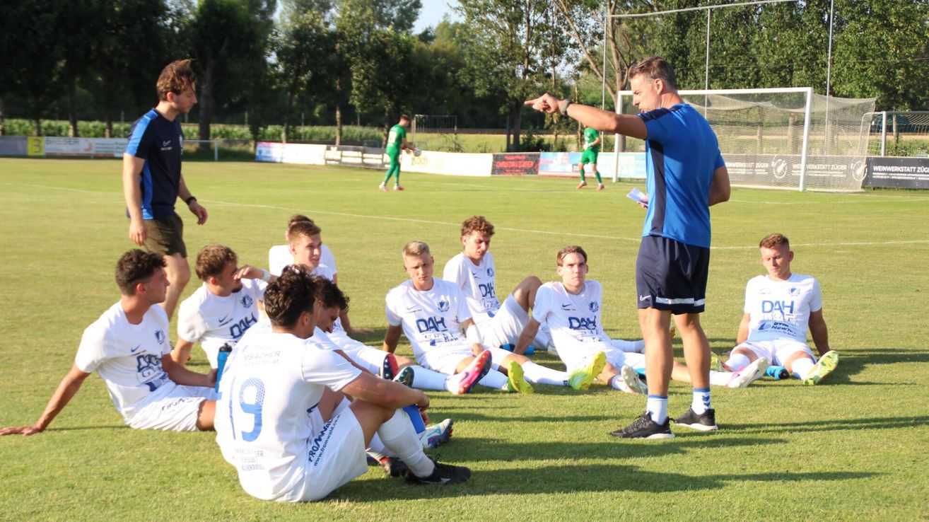 Ein Fußballtrainer zeigt auf das Spielfeld, während er sein Team anspricht, das auf dem Rasen sitzt. Spieler in weißen Trikots mit DAH lauschen aufmerksam. Im Hintergrund läuft ein Spieler in Grün auf das Tor zu.