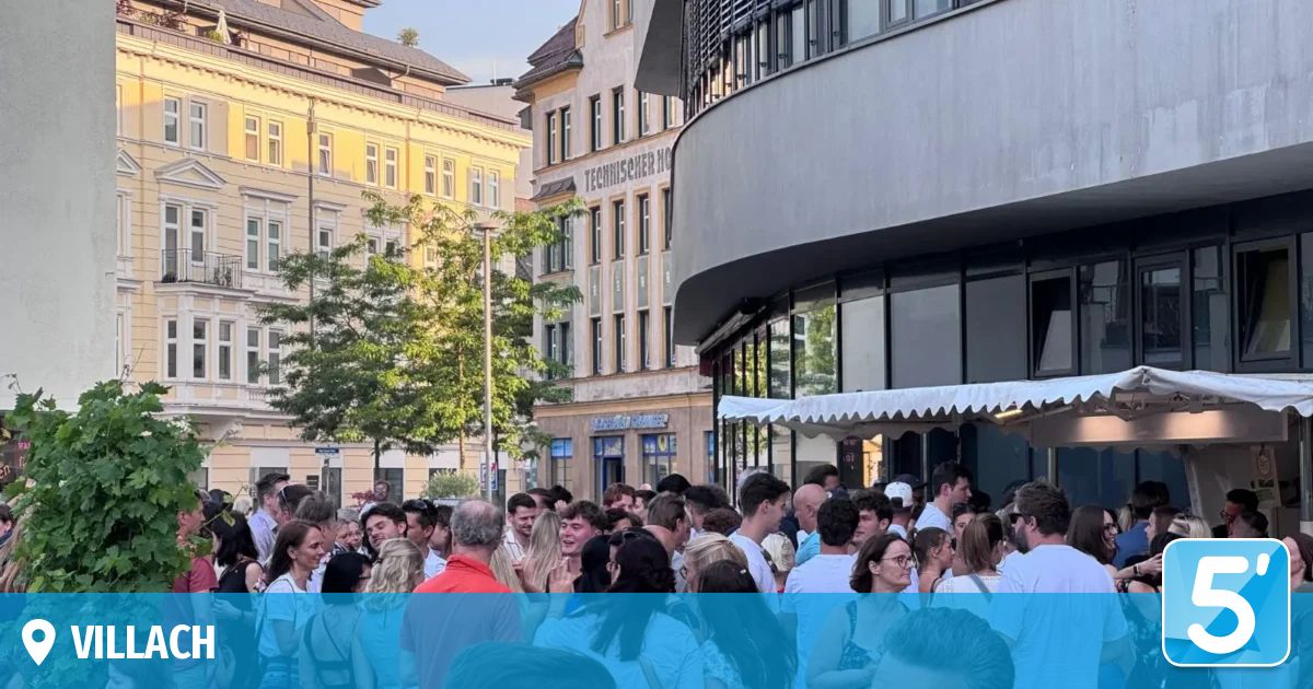 Eine Menschenmenge versammelt sich vor einem modernen Gebäude mit einem Baum im Vordergrund und einem gelben Gebäude im Hintergrund.
