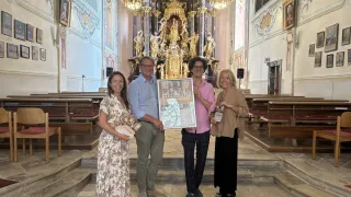 Vier Menschen stehen in einer Kirche, lächeln und halten ein gerahmtes Poster mit dem Text 'LIED DER SYRIA'. Dahinter befindet sich ein goldener Altar mit Statuen.