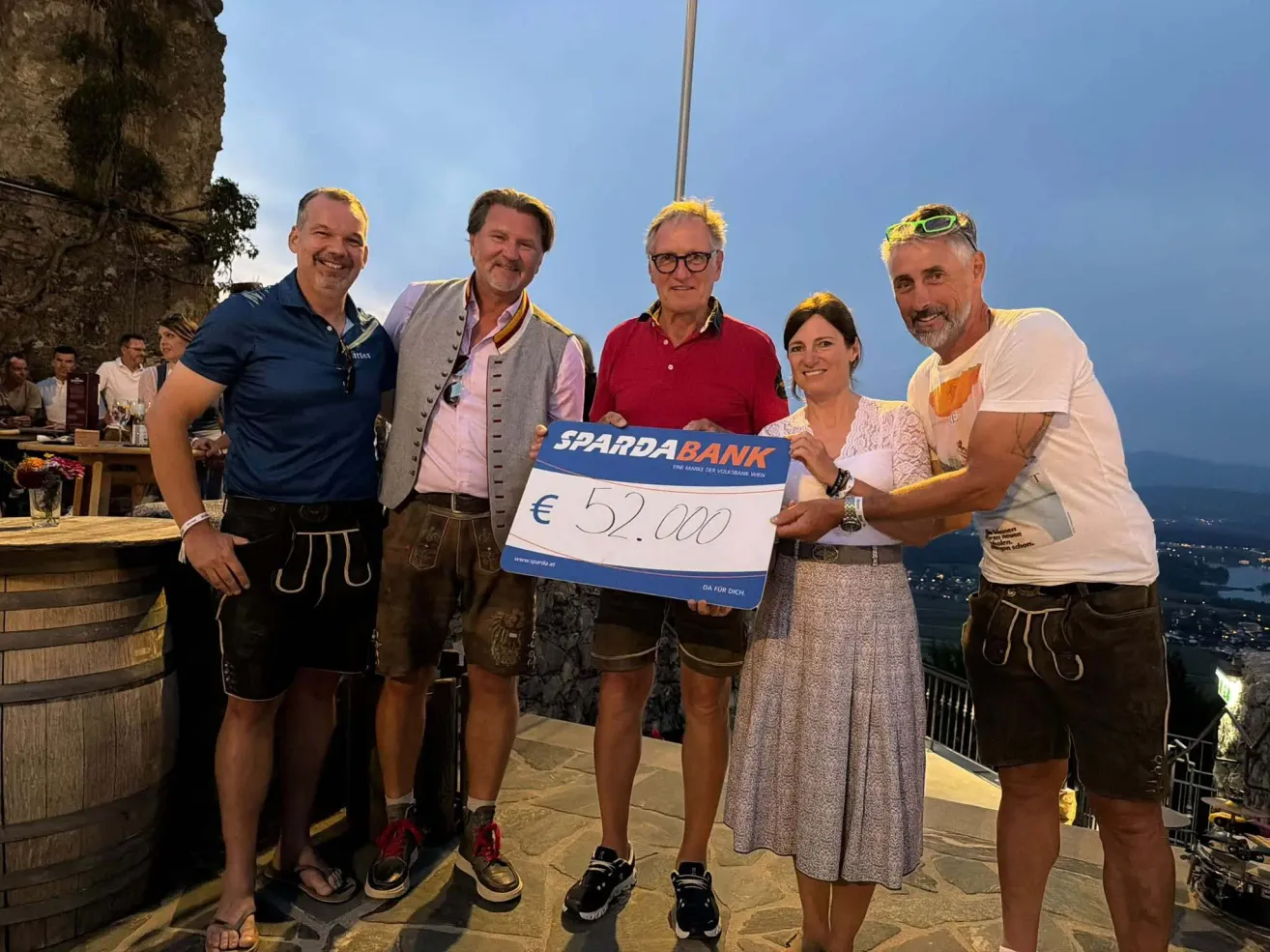 Fünf Menschen stehen zusammen und lächeln, halten einen großen blau-weißen Scheck über 52.000 Euro. Sie sind lässig gekleidet, einer trägt eine Brille und ein anderer eine Weste. Im Hintergrund ist eine landschaftliche Aussicht zu sehen.
