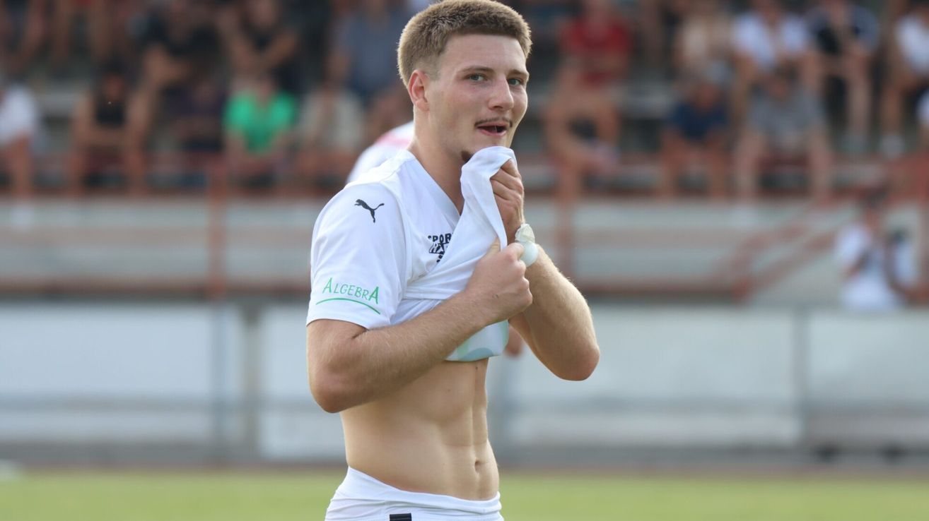 Ein Fußballspieler in einem weißen Trikot mit dem Wort Algebra darauf, das während eines Spiels Schweiß von seinem Gesicht wischt.