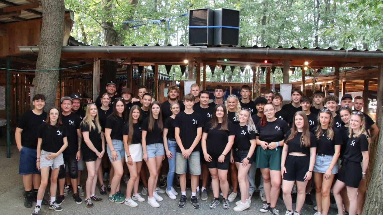A group of people wearing black t-shirts with white letters are posing for a photo in front of a wooden structure with speakers on top.