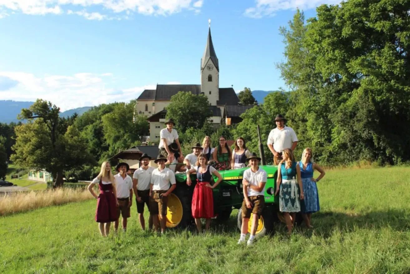 Eine Gruppe von Menschen in traditioneller Kleidung posiert vor einem Traktor mit einer Kirche im Hintergrund.