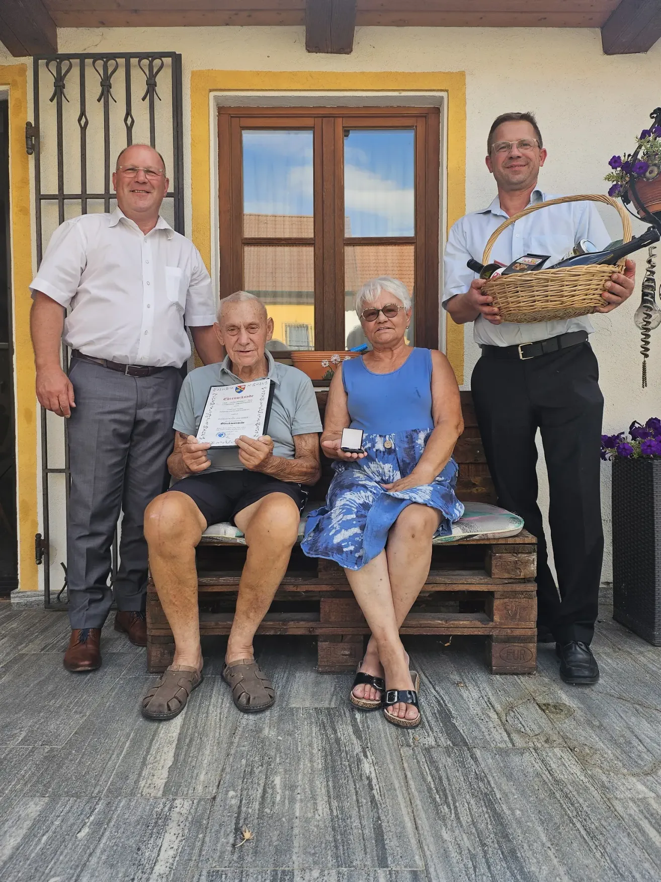 Vier Personen stehen auf einer Veranda, zwei von ihnen sitzen auf einer Holzbank. Der Mann auf der linken Seite hält ein Zertifikat und lächelt. Die Frau neben ihm hält ein kleines Objekt. Der Mann auf der rechten Seite hält einen Korb mit Flaschen. Der Mann, der hinter ihnen steht, lächelt.