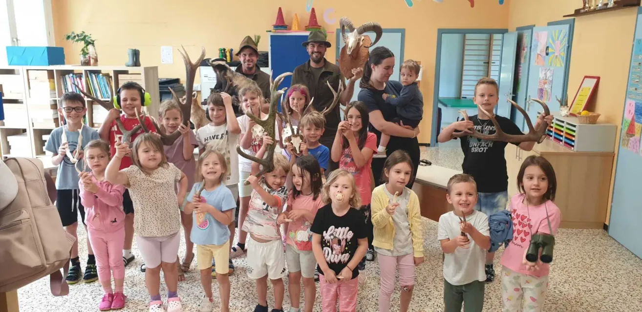 A group of children and adults are posing for a photo in a room with antlers and animal skulls on display. The children are holding antlers and some are wearing hats.
