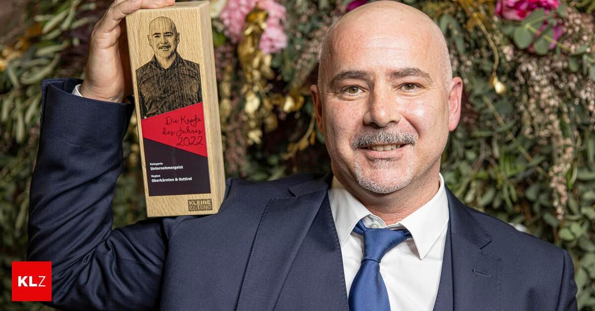 A man in a suit holding a wooden award that reads 'Die Koefe des Jahres 2022' with a picture of a man on it.
