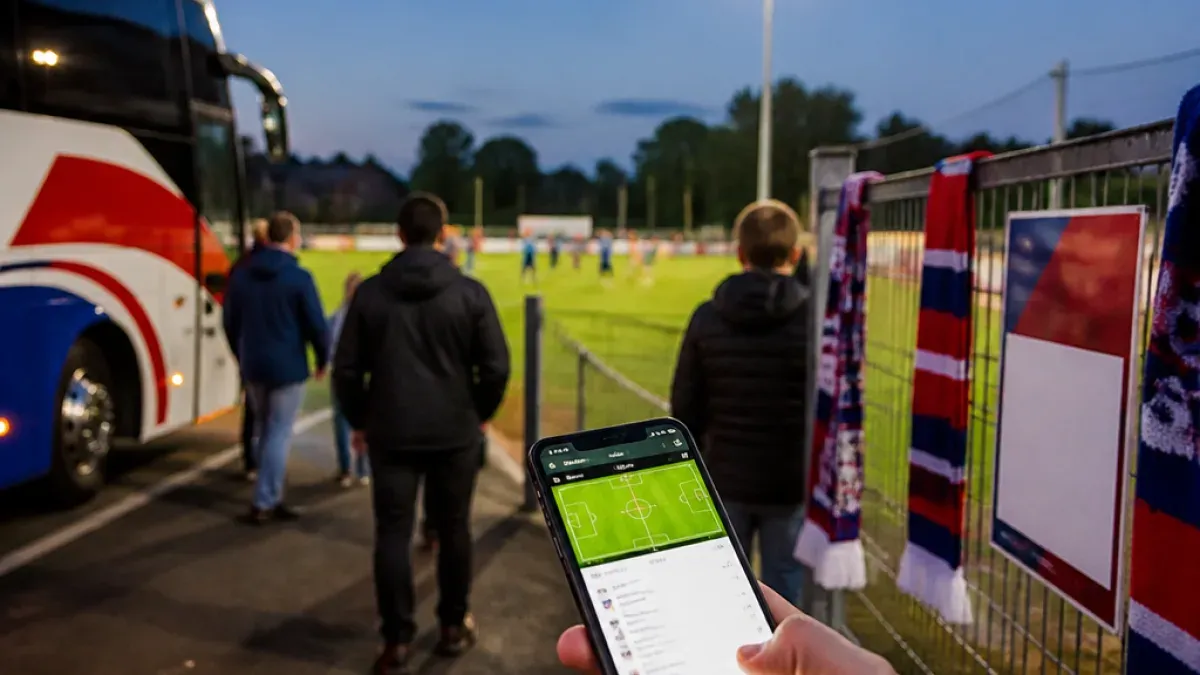 Eine Person, die ein Smartphone mit einem auf dem Bildschirm angezeigten Fußballspiel in der Hand hält, steht vor einem Zaun, an dem ein Schal hängt.