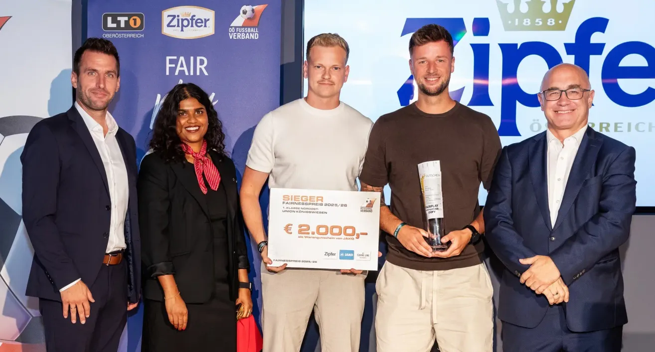 Three people stand together, holding a check and a trophy. The man in the middle holds a check for €2,000, while the man on the right holds a trophy. The woman on the left wears a red scarf. Behind them is a wall with logos.