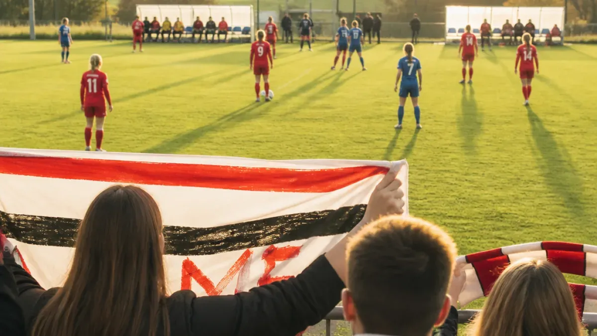 Ein Fußballspiel in vollem Gange mit Spielern in roten und blauen Uniformen, ein Spieler hält eine Fahne mit dem Wort 'ME' darauf.