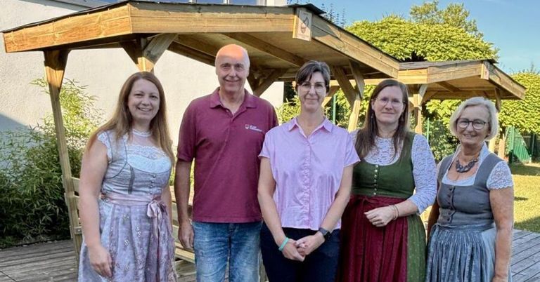 Five people are standing together in front of a wooden gazebo, smiling for a photo. The man in the middle wears a maroon shirt, while the women wear traditional dresses.