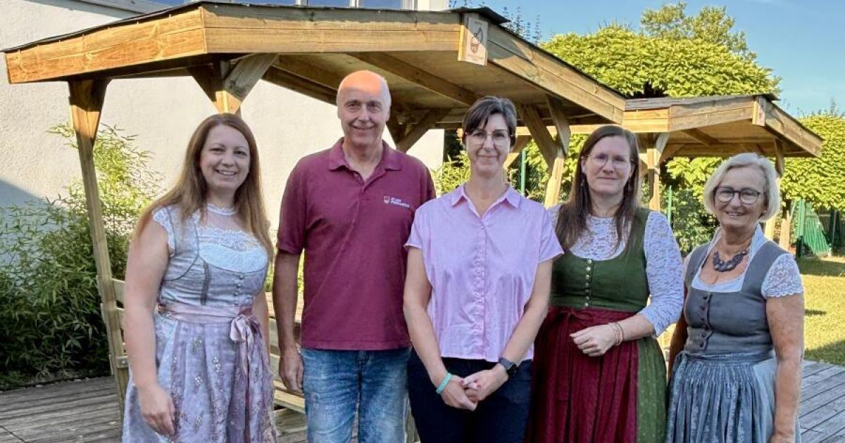 Five people are standing together in front of a wooden gazebo, smiling for a photo. The man in the middle wears a maroon shirt, while the women wear traditional dresses.