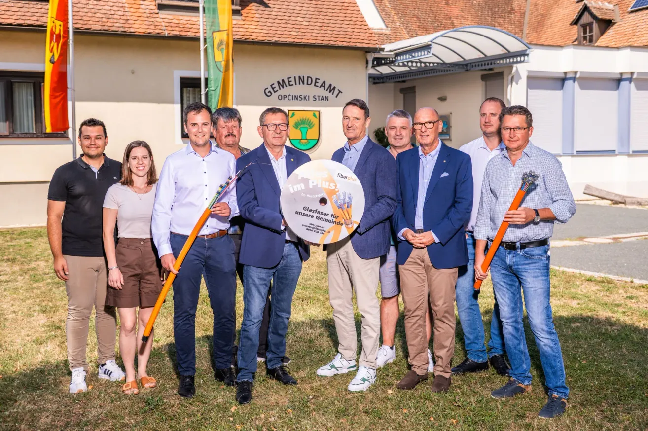 Eine Gruppe von Männern und Frauen steht vor einem Gebäude, hält ein großes kreisförmiges Objekt und lächelt für ein Foto.