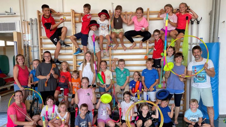 Eine Gruppe von Kindern und Jugendlichen posiert für ein Foto, während sie verschiedene bunte Requisiten wie Hula-Hoops, Pom-Poms und Jonglierstäbe halten. Sie stehen und sitzen auf einer Holzkletterwand.