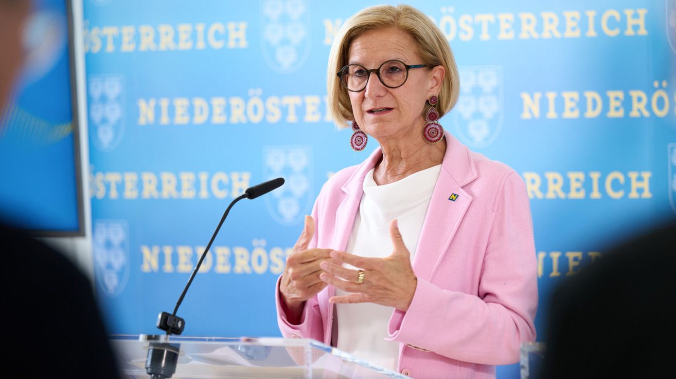 Eine ältere Frau mit Brille und einem rosa Blazer spricht an einem Podium mit einem Mikrofon vor einem blauen Hintergrund.