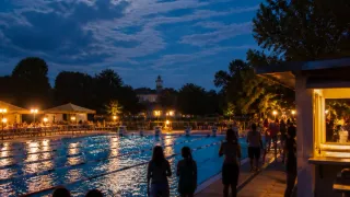 Eine Gruppe von Menschen geht nachts um einen Swimmingpool herum. Der Pool ist von Lichtern beleuchtet und im Hintergrund sind Bäume und Gebäude zu sehen.