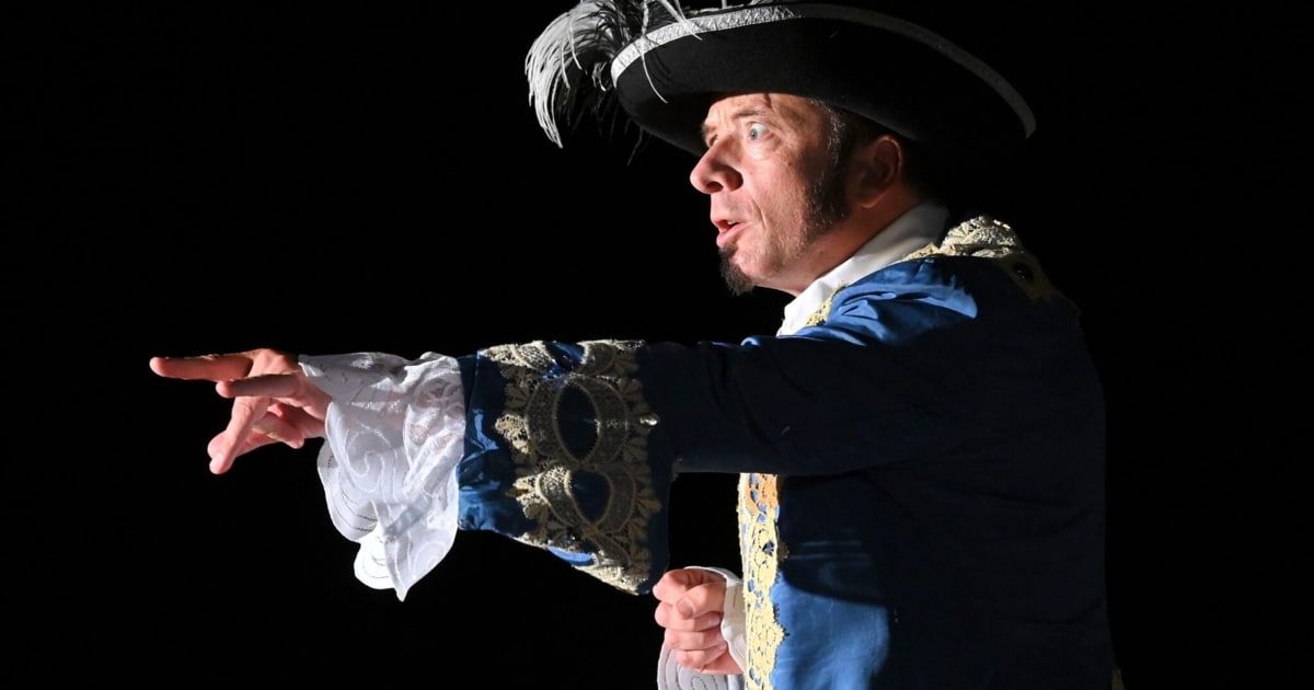 A man dressed in a historical costume with a black hat and a white feather points towards the left. He wears a blue and gold robe with intricate designs.