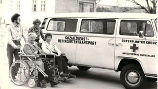 Ein Schwarz-Weiß-Foto von drei Menschen in Rollstühlen vor einem Van mit der Aufschrift 'Sozialdienst Behindertentransport'.