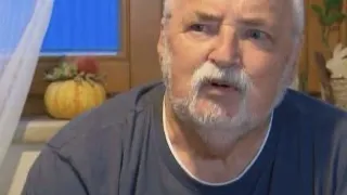 Ein älterer Mann mit weißem Bart und Schnurrbart sitzt vor einem Mikrofon und trägt ein dunkelblaues Hemd. Hinter ihm befindet sich ein Fenster mit einem weißen Vorhang und einem kleinen Kürbis auf der Fensterbank.
