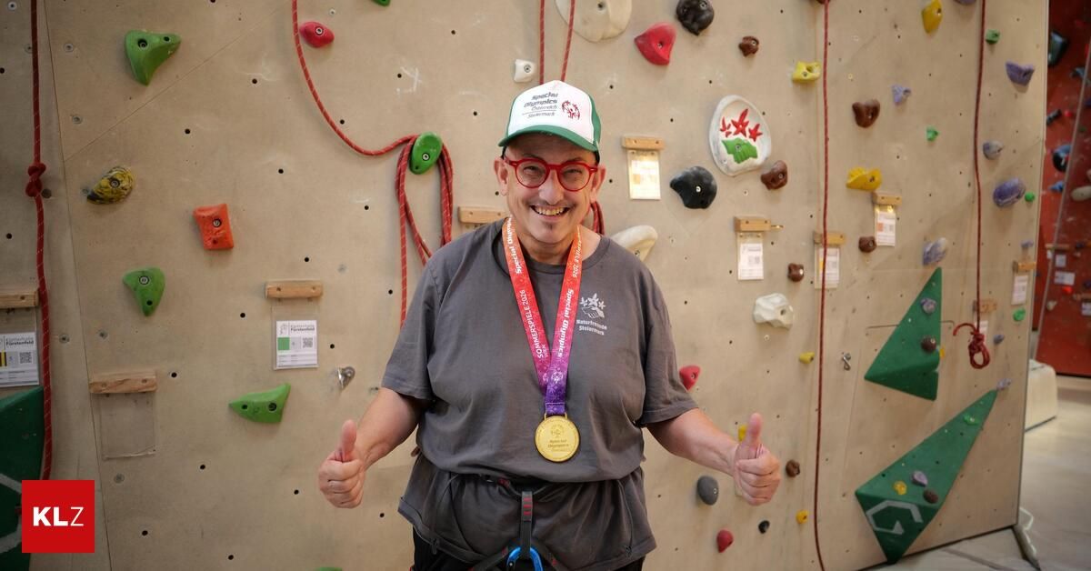 Ein Mann mit einer Kappe und Brille gibt ein Daumen hoch vor einer Kletterwand mit verschiedenen farbigen Griffen und einer Medaille um den Hals.