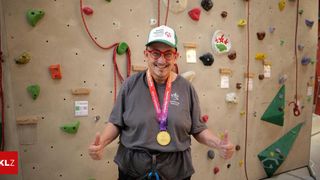 A man wearing a cap and glasses is giving a thumbs up in front of a rock climbing wall with various colored grips and a medal around his neck.
