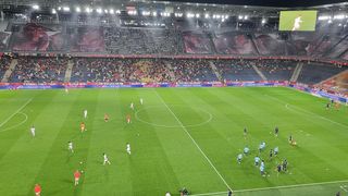 Ein Fußballspiel findet in einem Stadion mit einem großen Publikum statt. Spieler in roten und blauen Uniformen sind über das Feld verteilt. Das Stadion ist gut beleuchtet mit hellen Lichtern, und Werbung ist auf den Tribünen sichtbar.