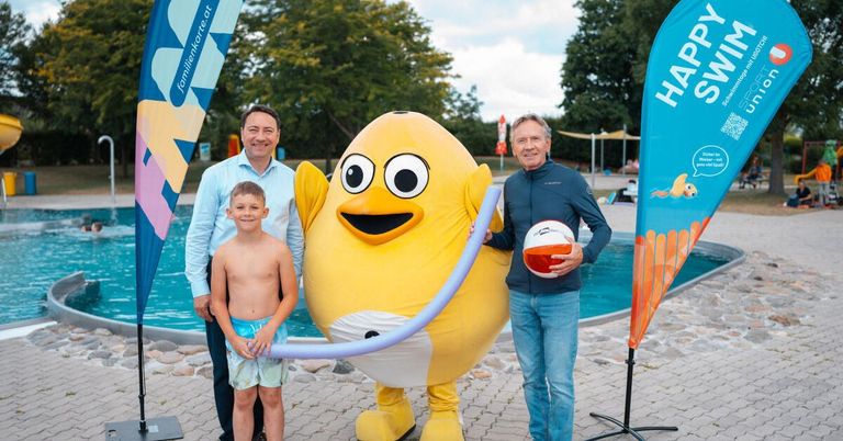 Three individuals, including a boy in swim trunks, stand beside a large yellow mascot holding a beach ball, with a swimming pool and banners in the background.