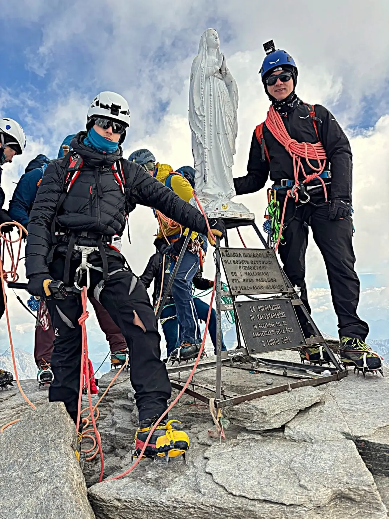 Eine Gruppe von Kletterern in Winterausrüstung steht auf einem Berggipfel, wobei ein Kletterer ein Seil hält und ein anderer neben einer Statue steht.