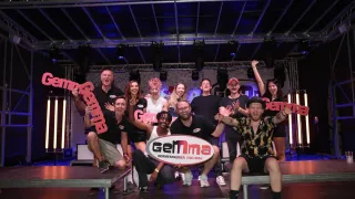 Eine Gruppe von Menschen auf einer Bühne, die lächeln und für ein Foto posieren. Sie tragen schwarze T-Shirts mit dem Wort 'GemMa' darauf. Dahinter befindet sich ein großes Banner mit demselben Wort.