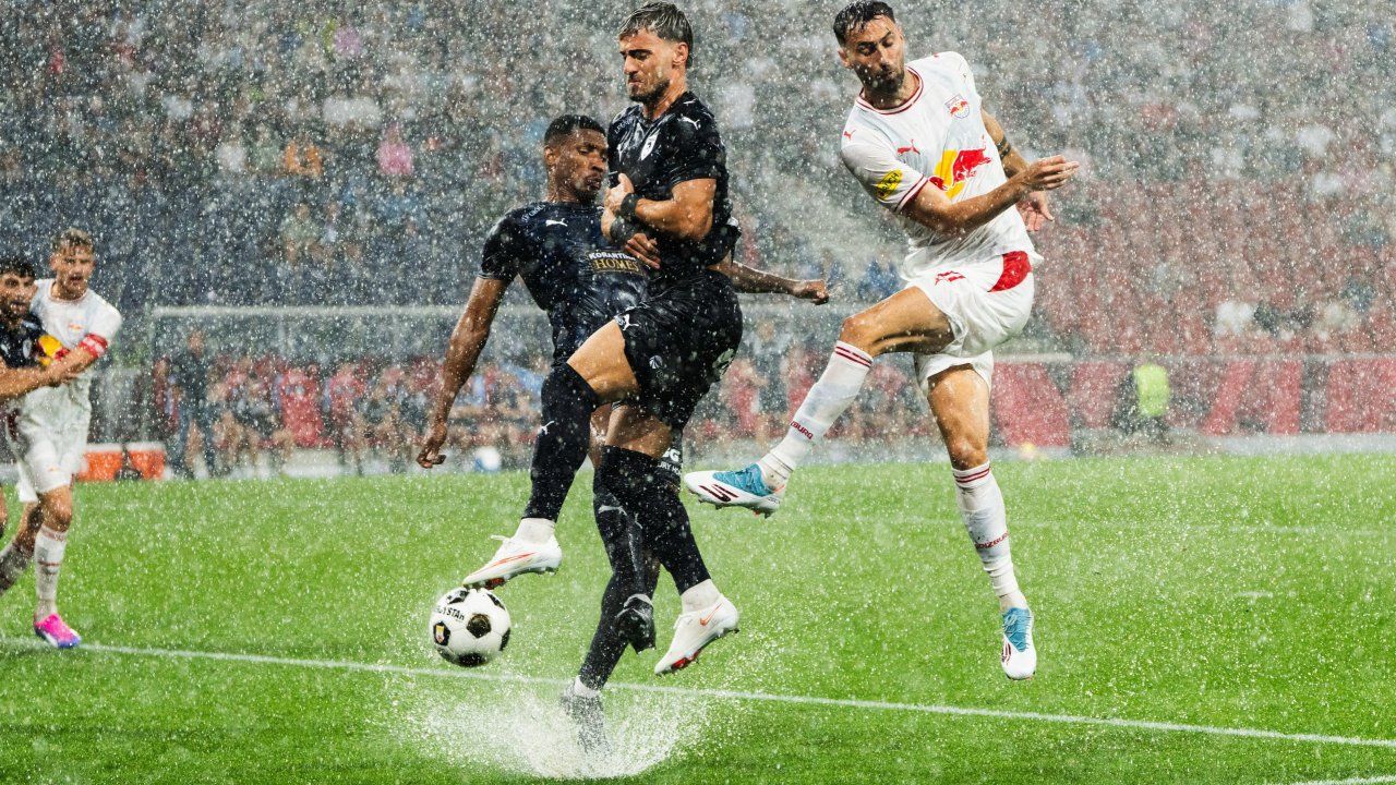 Drei Fußballspieler in Aktion während eines Spiels im Regen, einer in Schwarz und zwei in Weiß.