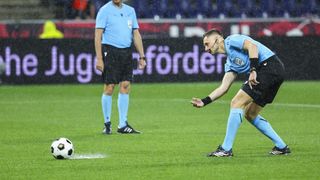 Ein Fußballschiedsrichter beugt sich nach unten, um den Boden zu inspizieren, während ein anderer Schiedsrichter hinter ihm steht. Das Feld ist nass, und ein Fußball liegt auf dem Boden.