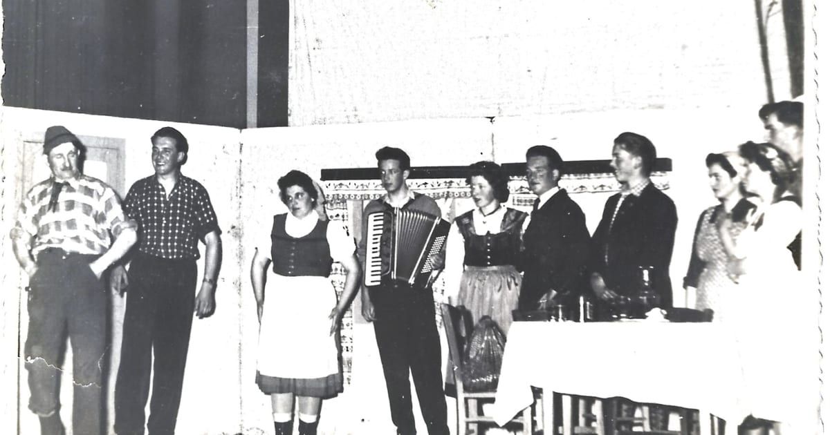 Ein Schwarz-Weiß-Foto zeigt eine Gruppe von Menschen in traditioneller Kleidung, die vor einer Wand mit einem gemusterten Design stehen. Ein Mann spielt Akkordeon, während andere zusehen.